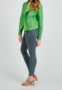 Donna che indossa una giacca verde brillante con zip, jeans skinny grigi e sandali con tacco alto color sabbia, in piedi contro uno sfondo chiaro e semplice.