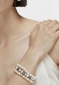 Pulsera de perlas en capas que presenta perlas redondas y en forma de óvalo en tonos blanco, crema y oscuros, con acentos dorados, descansando sobre un brazo suave.