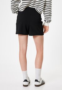 Schwarze, taillierte Shorts mit glatter Textur und zwei Seitentaschen. Getragen mit weißen Knöchelsocken und schwarz-weißen Sneakers. Gestreiftes Oberteil sichtbar.
