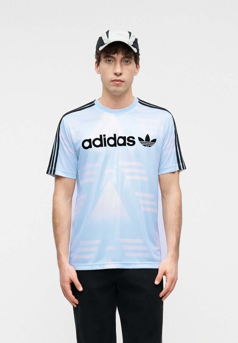 Junger Mann, der ein hellblaues Adidas-T-Shirt mit schwarzen Streifen an den Ärmeln und eine schwarz-weiße Mütze trägt, steht vor einem schlichten Hintergrund.