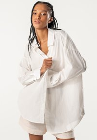White oversized shirt made of smooth cotton, featuring a classic collar, long sleeves, and button closure, with a curved hem.