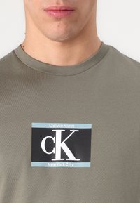 Olivovo zelené bavlnené tričko s okrúhlym výstrihom, ktoré má v strede potlač loga v čiernej a bielej farbe s nápismi "Calvin Klein" a "New York City".