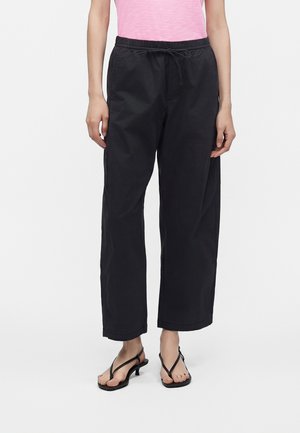 Persona che indossa un top rosa, pantaloni neri larghi con coulisse e sandali neri con tacco, in piedi su uno sfondo bianco semplice.