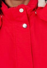 Chaqueta impermeable roja con un cuello alto, botones a presión plateados y textura acolchada en el interior. Detalles de material esponjoso visibles.