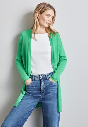 Maglione verde con maniche lunghe, apertura frontale e polsini a coste, indossato sopra una maglietta bianca e abbinato a jeans blu a vita alta.