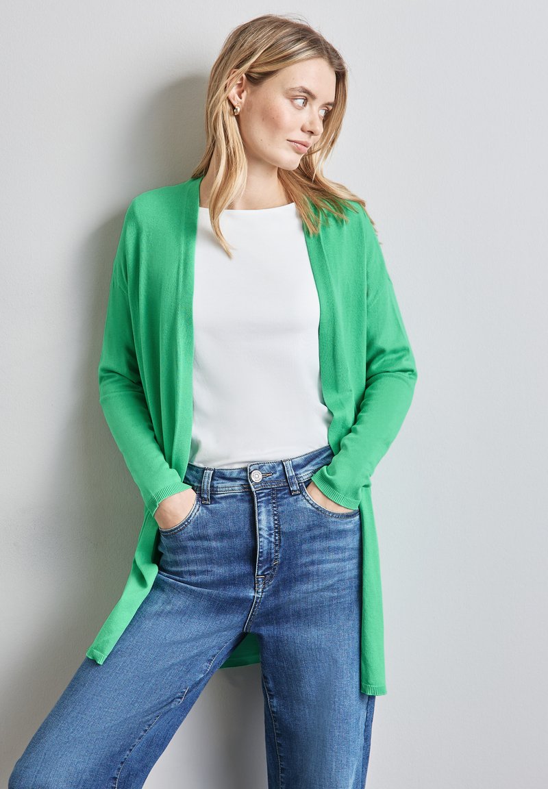 Grön kardigan med långa ärmar, öppen front och ribbade muddärmar, bärs över en vit topp och kombineras med blå jeans med hög midja.