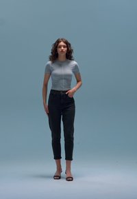 Κοντομάνικο ανοιχτό μπλε τοπ συνδυασμένο με σκούρο τζιν και μαύρα σανδάλια. Το μοντέλο στέκεται όρθιο μπροστά σε ανοιχτό μπλε φόντο.