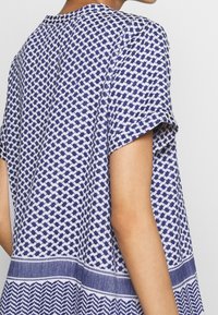 Blå och vit mönstrad topp med korta ärmar, som har en geometrisk design och texturerad tyg. Sömmen har en kontrasterande randig kant.