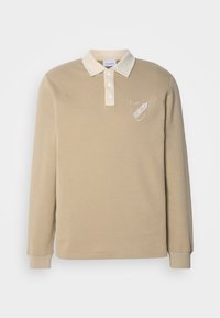 Cămașă polo cu mâneci lungi de culoare bej, având guler crem, cu o plăcuță cu patru butoane și un emblema brodată pe partea stângă a pieptului. Textură moale.