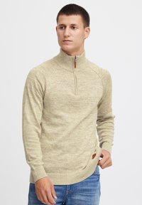 Vaaleanbeige neulepusero, jossa on korkea kaulus, puolivetoketju nahkavetimellä, ribattu hihansuu ja sivussa logo-ompelu. Slim fit -muotoilu.