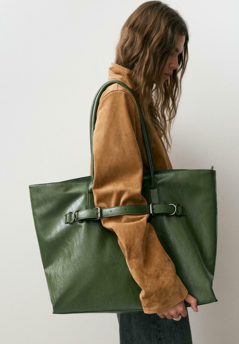 Vrouw met bruine suède jas draagt een grote groene leren shopper over haar schouder tegen een effen achtergrond.