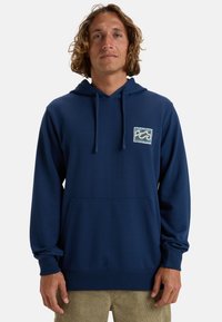 Sudadera azul marino hecha de tela suave, con un bolsillo delantero y un logo de ola bordado en el pecho. Capucha con cordón ajustable.