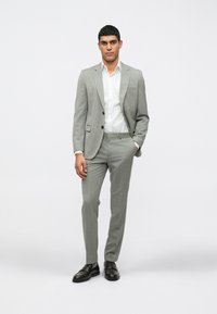 Ανοιχτό γκρίζο κοστούμι από υφή υφάσματος, με σακάκι ενός κουμπί και παντελόνι slim-fit. Συνδυασμένο με λευκό πουκάμισο και μαύρα παπούτσια.