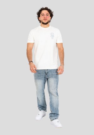 Joven con cabello rizado que lleva una camiseta blanca con un pequeño dibujo animado, vaqueros azul claro y zapatillas blancas, de pie sobre un fondo neutro.
