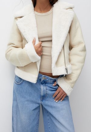 Femme portant une veste en peau lainée crème avec une fermeture éclair argentée, un haut côtelé beige et un jean ample taille haute bleu clair, posant les mains près de la taille.