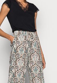 T-shirt noir avec bordure en dentelle au niveau de l'encolure, associé à un pantalon large à motifs avec des tourbillons et des designs floraux en beige, sarcelle et noir.