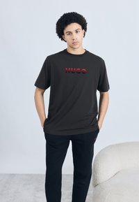 Čierne tričko s krátkym rukávom s plastickým červeným logom "HUGO" na prednej strane, doplnené čiernymi textúrovanými nohavicami. Minimalistický dizajn, voľný strih.