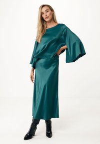 Abito in satin turchese con maniche svasate, vestibilità rilassata e gonna lunga. Abbinato a stivali neri con tacco, su uno sfondo neutro.