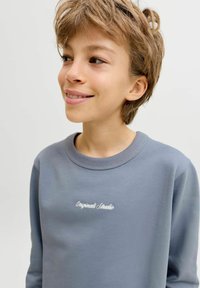 Χαμογελαστό παιδί με κοντά ανοιχτό καστανά μαλλιά που φοράει μπλε μπλούζα με κέντημα "Originals Studio" στο στήθος, κοιτάζοντας προς τα αριστερά.