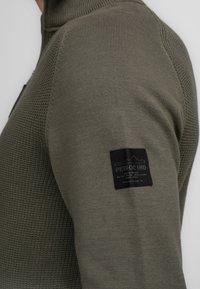 Olivgrüner Strickpullover mit hohem Kragen, ausgestattet mit strukturierten Mustern und einem schwarzen Etikett am Ärmel. Glatte Stoffqualität mit klaren Nähten.