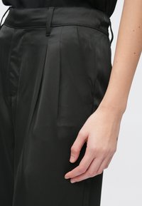 Czarne spodnie z satynowej tkaniny z przednimi zakładkami, gładką fakturą i dopasowanym pasem, pokazane z ręką opartą na boku.