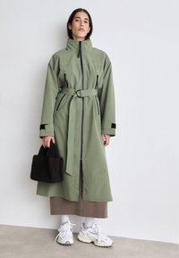 Cappotto lungo con cintura in tessuto verde oliva con accenti neri, tasche con zip e colletto alto. Abbinato a una borsa nera dalla texture pelosa e a scarpe da ginnastica bianche.
