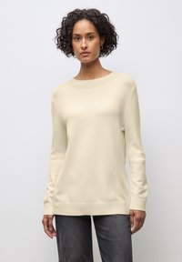 Donna con capelli corti e ricci che indossa un maglione crema a maniche lunghe e pantaloni grigio scuro, in piedi contro uno sfondo semplice grigio chiaro.