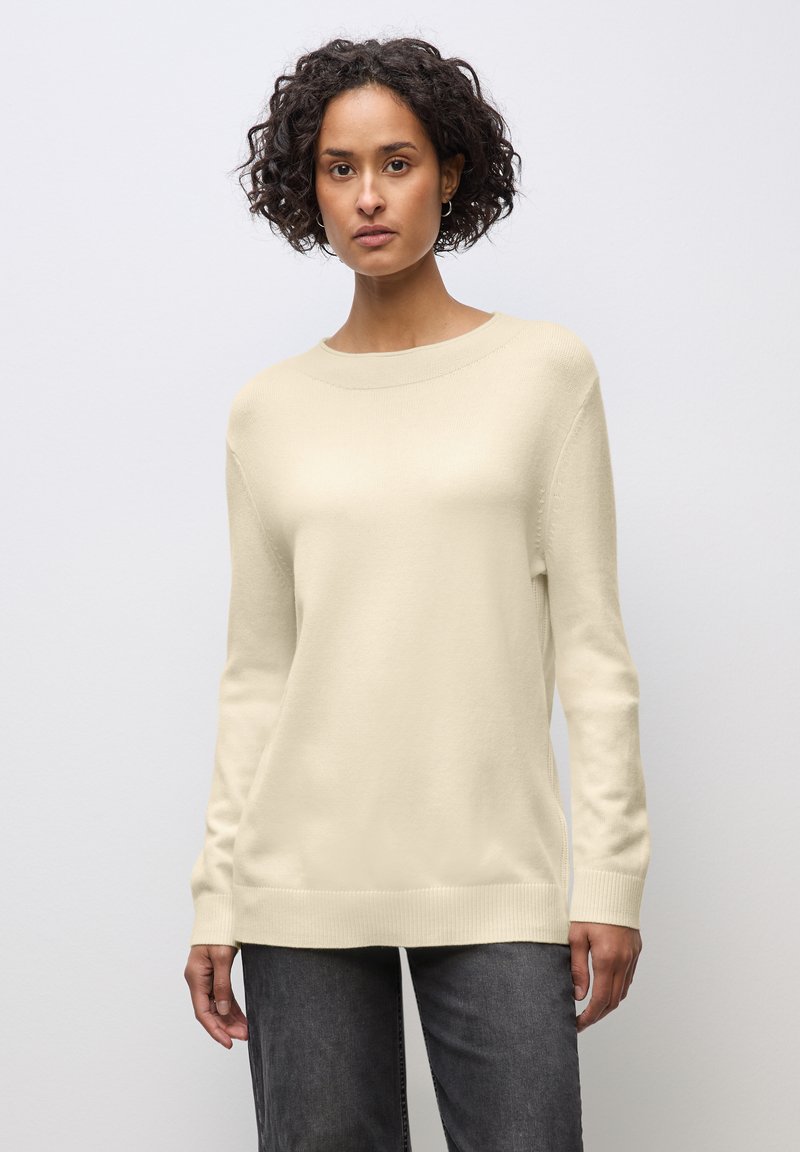 Donna con capelli corti e ricci che indossa un maglione crema a maniche lunghe e pantaloni grigio scuro, in piedi contro uno sfondo semplice grigio chiaro.