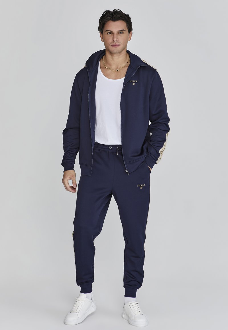 Chándal navy con acentos dorados, chaqueta con cremallera y capucha, puños elásticos, combinado con pantalones a juego y zapatillas blancas.