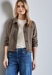 Brun knappad jacka med krage, ribbade muddar och en mjuk textur, bärs över en vit skjorta och kombineras med blå jeans.