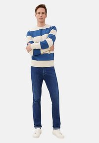 Suéter a rayas azules y blancas, diseño entallado, combinado con vaqueros azul oscuro y zapatillas blancas. Modelo parado con los brazos cruzados, fondo neutro.