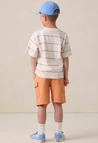 Enfant portant un t-shirt à manches courtes crème à rayures et un short cargo orange, une casquette bleue et des baskets à carreaux avec des chaussettes blanches.