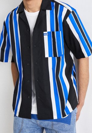 Man wearing black, blue, and white vertical striped short-sleeve button-up shirt over white t-shirt with light blue jeans.