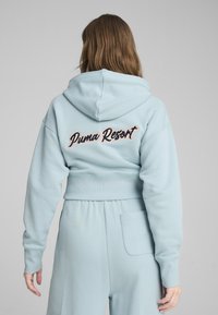 Svetlomodrý cropped mikina s kapucňou a rebrovaným lemom, so zafírovaným nápisom "Puma Resort" v kontrastných farbách na zadnej strane.