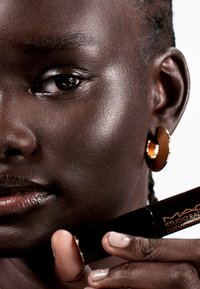 Tubo de maquillaje cilíndrico negro sostenido por una mano con uñas rosa claras, con acentos en dorado. El modelo lleva pendientes de aro dorados. Textura de piel suave.