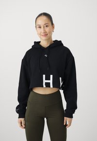 Hoodie cropped nero con grafiche bianche, cappuccio con cordini e orlo a coste; abbinato a leggings verde oliva a vita alta.