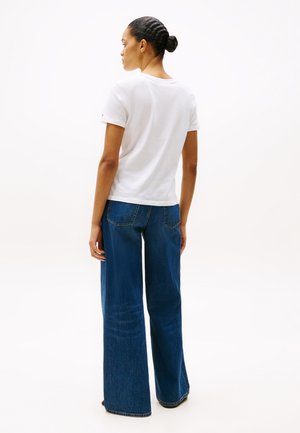 Kobieta z zaplecionym koczkiem, ubrana w zwykły biały t-shirt i szerokie niebieskie dżinsy, stojąca tyłem na białym tle.