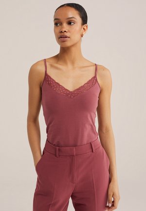 Mujer con camisola color malva con encaje y pantalones de cintura alta a juego, de pie con una mano en el bolsillo, fondo neutro.