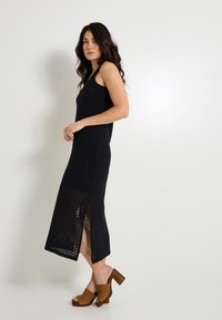 Vestido maxi de punto de ganchillo negro con aberturas laterales y un corte holgado. Combinado con sandalias de tacón grueso marrones con tachuelas metálicas.