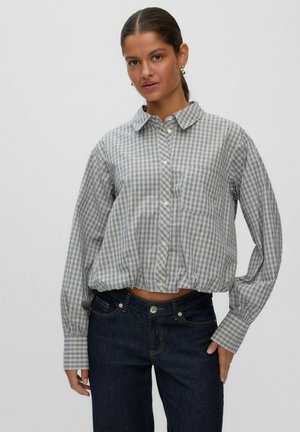 Donna con capelli scuri raccolti in una coda bassa, orecchini d’oro, camicia a quadri abbottonata con vita stretta e jeans blu scuro su uno sfondo uniforme.