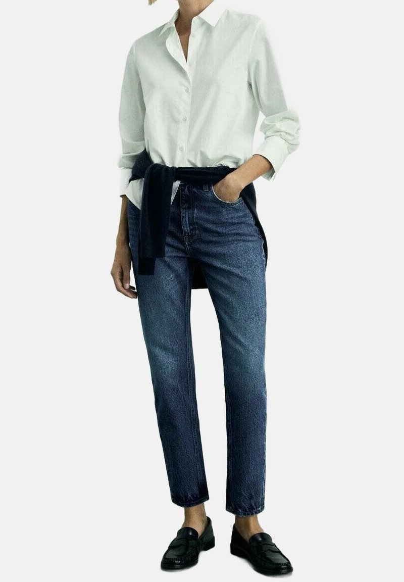 Chemise blanche boutonnée, jean bleu foncé, et mocassins noirs. La chemise a des manches retroussées, le jean est droit avec une légère décoloration.