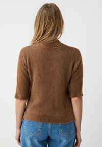 Bruine harige trui met korte mouwen en een ronde halslijn. Zachte textuur, lichtgewicht materiaal en een iets cropped ontwerp. Blauwe jeans zichtbaar daaronder.