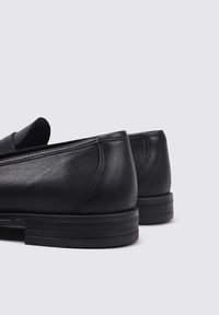 Mocasines de cuero negro con una textura suave, punteras redondeadas y una suela de goma plana con sutiles patrones de relieve. Acentos de costura simples.