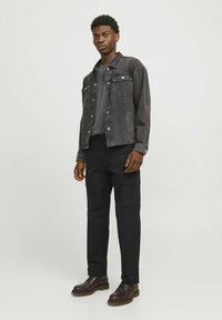 Sort denimjakke med to brystlommer, grå sweatshirts under, sorte cargo bukser og brune snørebåndsstøvler. Glat tekstur.