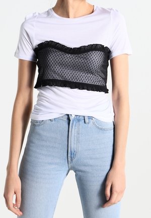 Žena nosí bílý krátký rukáv vrstvený s černým průsvitným volánkovým bandeau topem a světle modré džíny s vysokým pasem, stojící před jednobarevným pozadím.