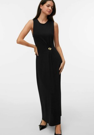 Žena oblečená v čiernych, bezrukávových maxi šatách so zlatou dekoratívnou sponou v páse, v kombinácii s čiernymi špicatými podpätkami, stojaca na bielom pozadí.