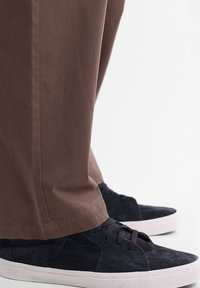 Baskets en daim noir avec semelle en caoutchouc blanche, associées à un pantalon en tissu marron, légèrement fuselé au niveau de la cheville, mettant en valeur la texture du matériau.