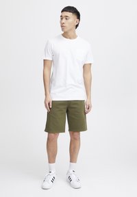 Vit bomullströja med bröstficka, tillsammans med olivgröna shorts. Modellen har på sig vita sneakers med svarta ränder och vita strumpor.