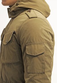 Homme portant une veste à capuche marron avec des poches à rabat boutonnées sur la poitrine et le haut du bras, vu de profil.