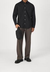 Man in een zwart blouse met knopen, bruine wijde broek, zwarte laarzen, die aan de hand een klein zwart ritszakje vasthoudt.